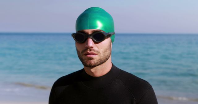 Depicts a confident male swimmer wearing a green swim cap and goggles standing on a beach with the ocean in the background. Ideal for use in campaigns related to sports, swimming, outdoor activities, and fitness, as well as promotional materials for swimwear brands and athletic gear companies.