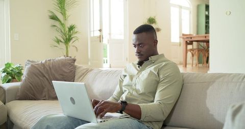 African american man working on laptop on bright sofa in open-plan living room