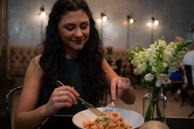 Woman enjoying a plate of pasta at a cozy restaurant. She is smiling and appears to be having a pleasant meal. A vase with flowers is on the table, adding to the warm ambiance. Ideal for use in lifestyle blogs, restaurant promotions, food-related advertisements, and social media content highlighting dining experiences.