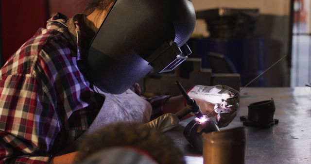 Depicts welder wearing protective gear working intensely on metal components in workshop setting. Ideal for illustrating manual labor, precision tasks, industrial workplace safety, and engineering professions. It highlights important aspects of trade skills and safety measures.