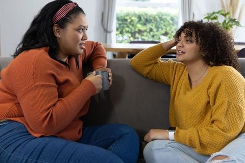 Two women chatting and relaxing on sofa with coffee mug, cozy casual home conversation