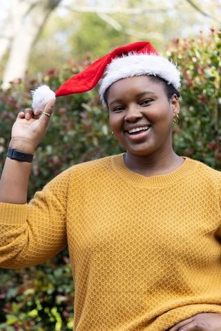 African american woman smiling holding santa hat pompom outdoors wearing mustard sweater
