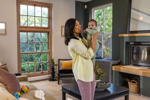 Smiling mother holding infant, baby reaching mouth in sunlit modern living room, cozy home