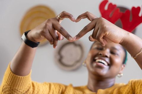 Smiling black woman making heart gesture wearing red antlers and mustard sweater in cozy home