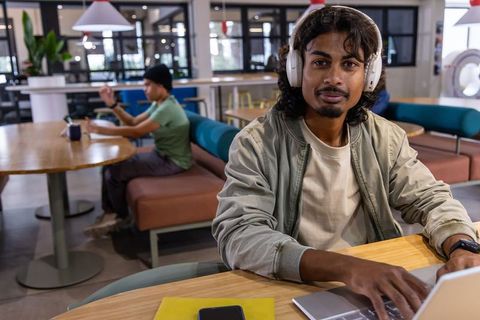 Indian man wearing headphones working on laptop in coworking lounge focused on productivity