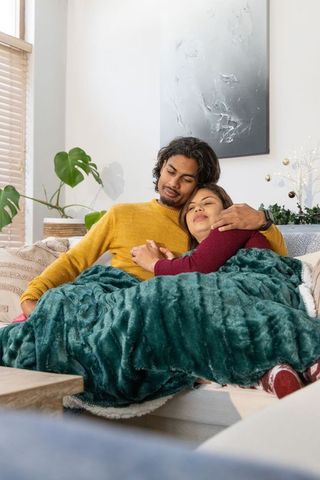 Asian couple cuddling on light sofa under teal plush blanket, cozy home romance and warmth