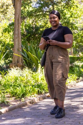 African american woman standing in sunlit park holding smartphone and smiling