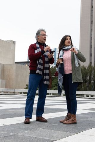 Mature couple navigating urban plaza, man pointing at smartphone, woman holding coffee