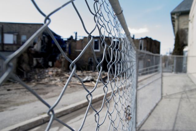 Chain-link fence focuses in foreground while background shows abandoned, decaying buildings. Useful for themes such as urban decay, neglect, environment, city decline, and socio-economic issues.