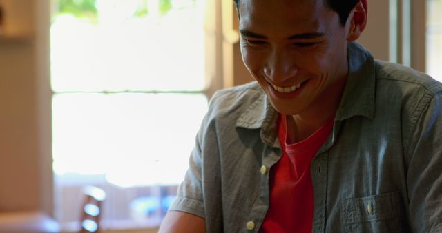 Smiling young Asian man wearing casual outfit, enjoying a moment indoors with natural light. Perfect for promoting positive mood, happiness, casual wear, and indoor activities.