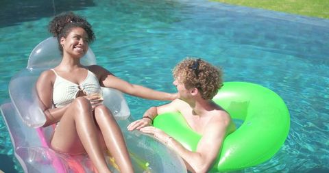 Mixed-race couple relaxing on inflatables in sunlit backyard pool