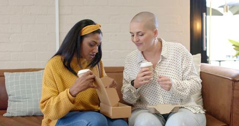 Diverse women sharing takeout and coffee on couch, enjoying casual cozy friendship moment