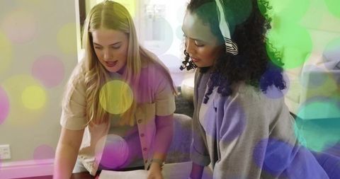 Two women collaborating reviewing documents in cozy living room with colorful bokeh