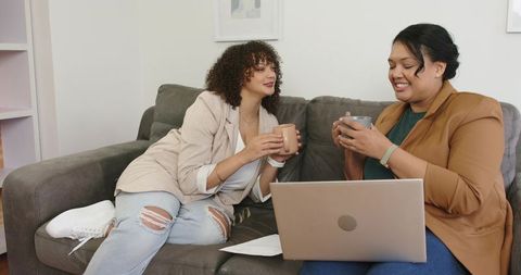 Two women sharing coffee and collaborating on laptop while relaxing on gray sofa in home office