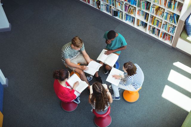 Overhead view of a group of diverse students studying together in a school library. They are sitting on colorful stools and reading books, indicating a collaborative learning environment. This image can be used for educational websites, school brochures, and articles about student life, teamwork, and academic collaboration.