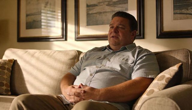 Middle-aged man sitting calmly on beige sofa clasping hands in sunlit living room