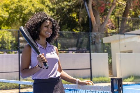 Smiling african american woman holding black paddle on blue court wearing lavender polo