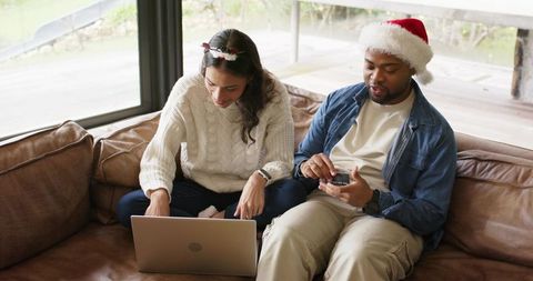 Festive diverse couple doing holiday online shopping on laptop with gift box and santa hat