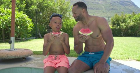 African american father and teen son enjoying watermelon by backyard pool on sunny day