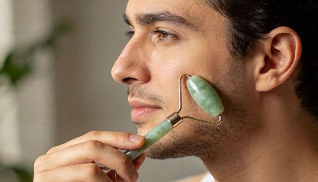 Closeup man using green jade roller along cheek for facial massage and grooming