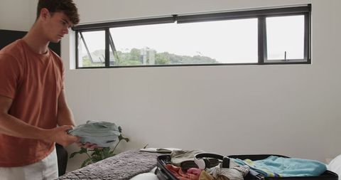 Young man packing luggage on bed in contemporary minimalist bedroom preparing for trip
