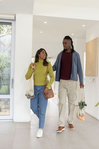 Multicultural couple walking through modern home carrying reusable grocery bags, smiling