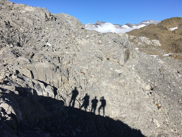Group of hikers casting shadows on rugged rocky terrain during a climb under a clear blue sky. Ideal for themes related to outdoor adventure, exploration, physical challenge, and group activities. Can be used in travel blogs, adventure magazines, motivational posters, and social media posts.