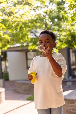 African american boy blowing soap bubbles on sunlit patio holding yellow cup