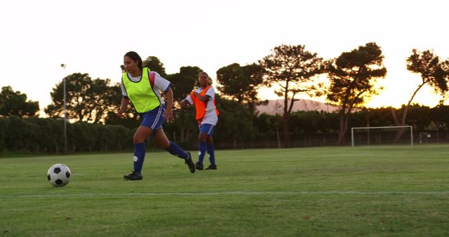 Two female soccer players are practicing on a well-maintained green field at sunset. One woman is in control of the ball while the other is in pursuit. The background features trees and an empty goalpost. This image can be used for promoting women's sports, athletic gear, sports training programs, and teamwork. It's ideal for articles or advertisements focusing on fitness, outdoor activities, and the spirit of competition.
