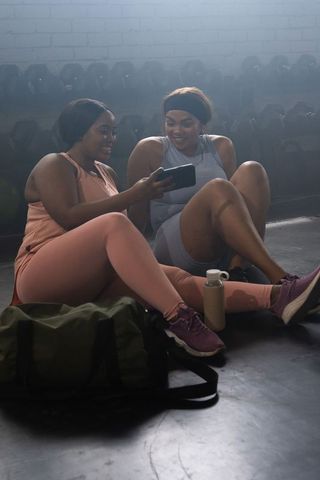 Two african american women resting, sharing smartphone and smiling after workout in gym