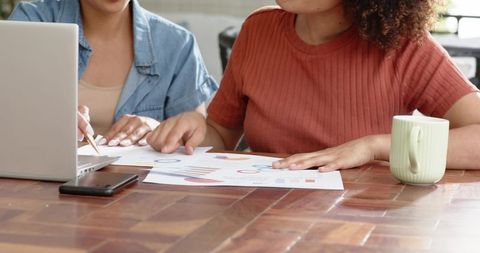 Multiracial women reviewing charts and collaborating on laptop for data analysis at home