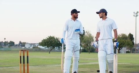 Two cricketers walking as batting partners on suburban cricket pitch at dusk carrying bats