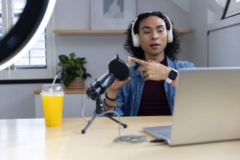 Non-binary creator recording podcast at home studio wearing headphones pointing at mic