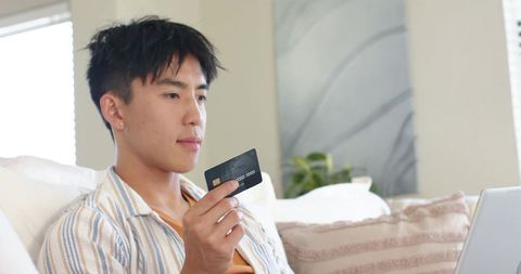 Young asian man holding credit card while shopping online on laptop at home