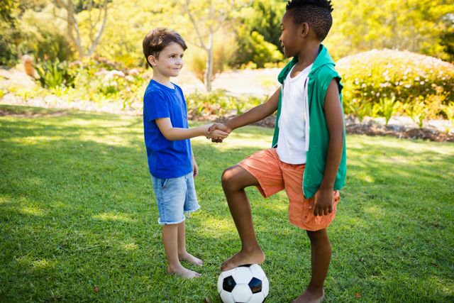Two boys are handshaking on a grassy soccer field in a park, symbolizing friendship and teamwork. One boy has his foot on a soccer ball, indicating a playful and sporty atmosphere. This image is ideal for promoting children's sports activities, teamwork, outdoor play, and summer fun.
