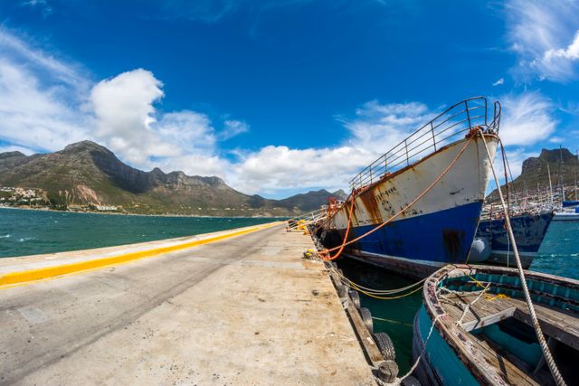 Rusty fishing boats docked at a coastal harbor under a bright blue sky with scenic mountain views in the background. Suitable for use in travel promotions, maritime industry ads, and scenic wallpaper.