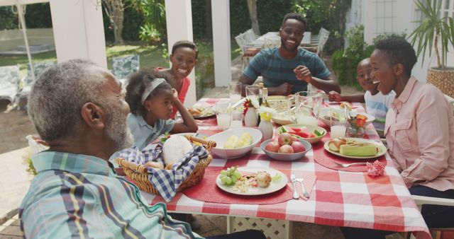 Happy african american family sitting at table in garden, eating dinner and talking. Lifestyle, domestic life, family, and togetherness.
