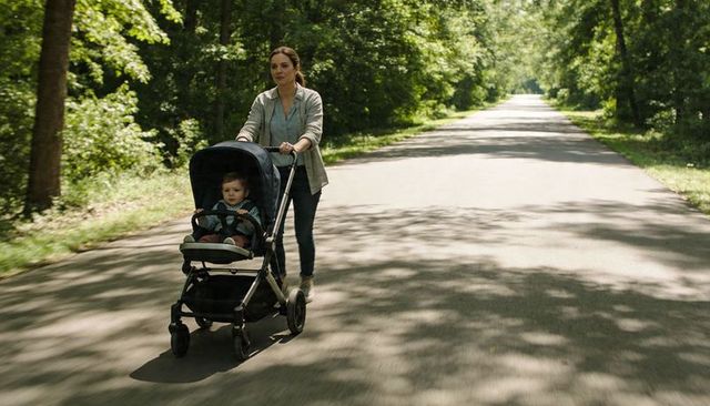 Young mother walking pushing stroller with toddler on sunlit tree-lined park road
