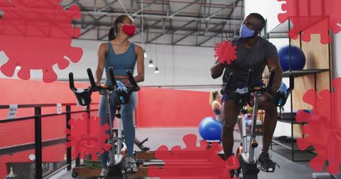 Masked fitness partners cycling on stationary bikes with red virus overlays in gym