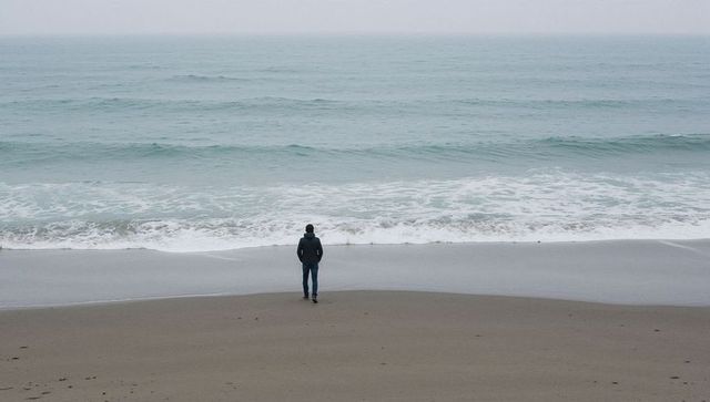 Solitary man standing on misty shoreline facing calm ocean waves for reflection