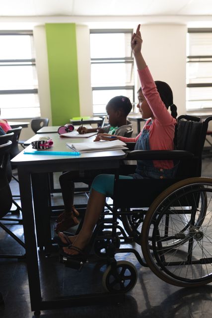 Young schoolgirl in a wheelchair actively participating in a classroom setting by raising her hand. Ideal for use in educational materials, diversity and inclusion campaigns, and articles promoting accessible education.