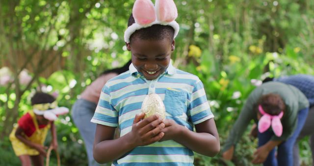 This image depicts children enthusiastically participating in an Easter egg hunt in a lush green garden. A boy is holding a sparkling golden egg, while the others are searching through the greenery, all wearing playful bunny ears. Ideal for illustrating spring holiday celebrations, family activities, and outdoor festivities in Easter-related content.