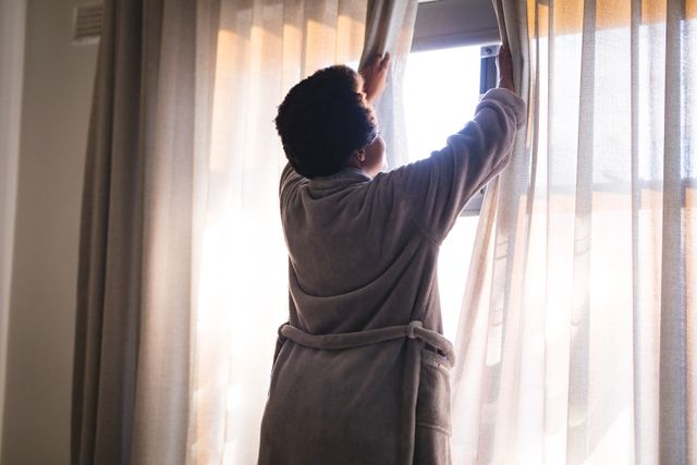 Rear view of african american mid adult woman opening curtains at window. unaltered, lifestyle, morning, waking up.