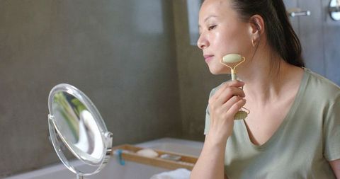 Asian woman using jade facial roller at bathtub mirror for natural skincare routine