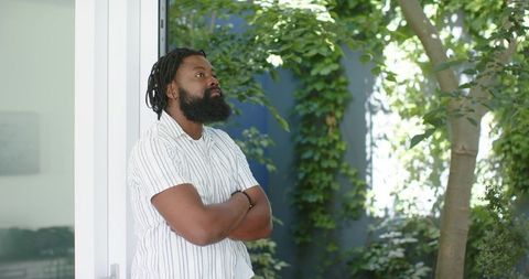 African american man standing in doorway gazing into sunlit courtyard draped in ivy and wearing stri