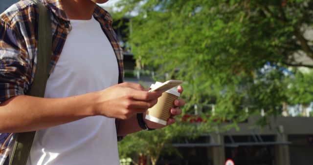 Young man wearing a casual shirt and backpack holding a coffee cup while checking his smartphone. Picture captures a typical modern lifestyle scene in an urban setting. Ideal for usage in technology, lifestyle, and urban living themed content, promoting mobile apps, coffee brands, or outdoor activities.