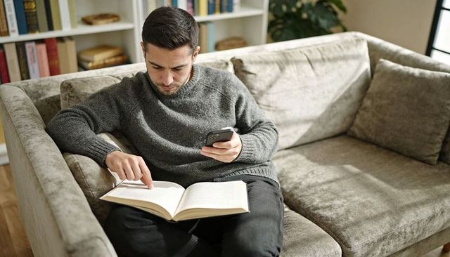 Man reading book on sofa while checking smartphone cozy home relaxation and multitasking