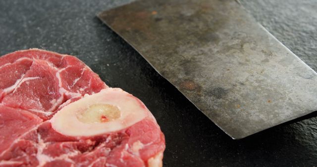 A raw beef shank cut with the bone in is placed next to a large cleaver on a dark surface, with copy space. The image showcases the ingredients and tools used in preparing meat dishes.