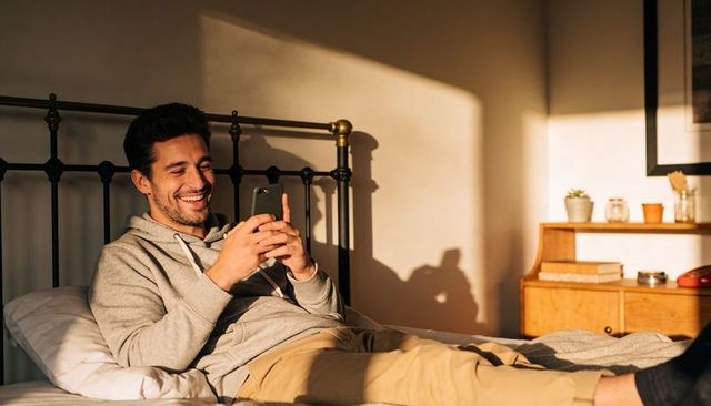 Man Relaxing on Bed Using Smartphone in Sunlit Bedroom with Metal Headboard