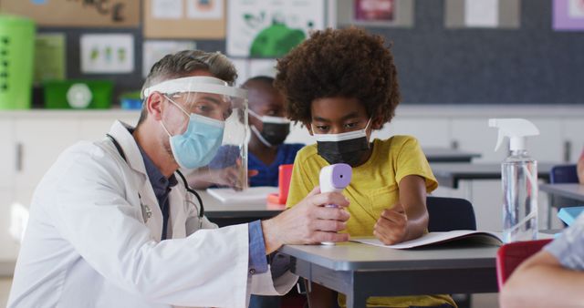 Healthcare professional wearing face shield and mask checking temperature of elementary student in the classroom. This image can be used for topics related to school safety, pandemic precautions, healthcare in education settings, and child wellness.